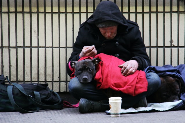 Picture of a homeless man sitting cross-legged on the ground curled over a dog in a red coat, on his lap. The dog is looking at the camera.
